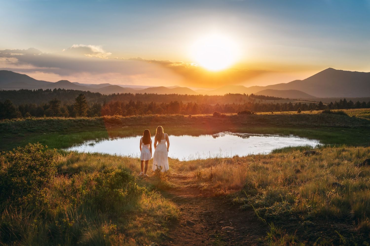 Two women in white dresses stand by a reflective pond watching a golden sunset over misty mountains.