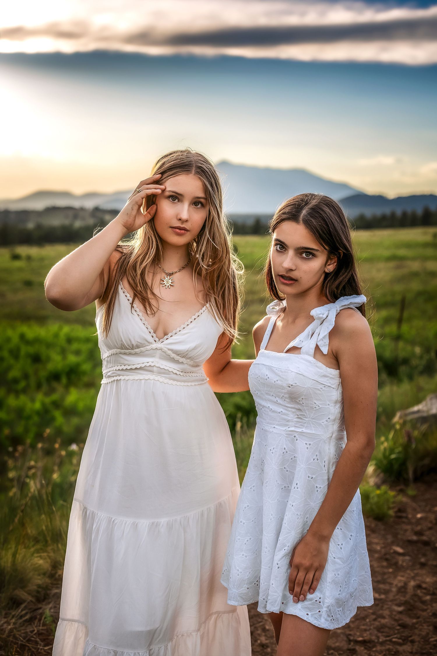 Two teenage girls in white dresses posing in a mountain meadow during a dramatic sunset.