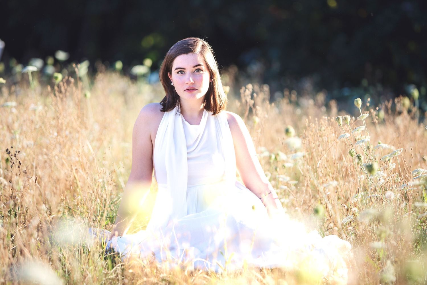 Young woman in white dress sitting peacefully in a sunlit golden meadow field.