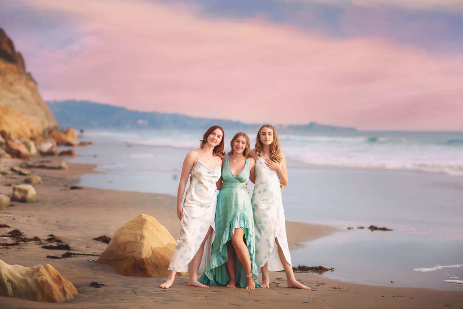 Three women in elegant dresses pose together on a sandy beach at sunset with pink sky.