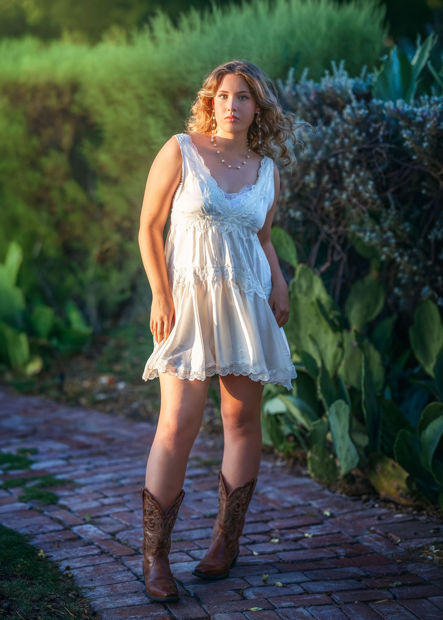 Young woman in white lace mini dress and brown cowboy boots standing on brick path surrounded by lush greenery.