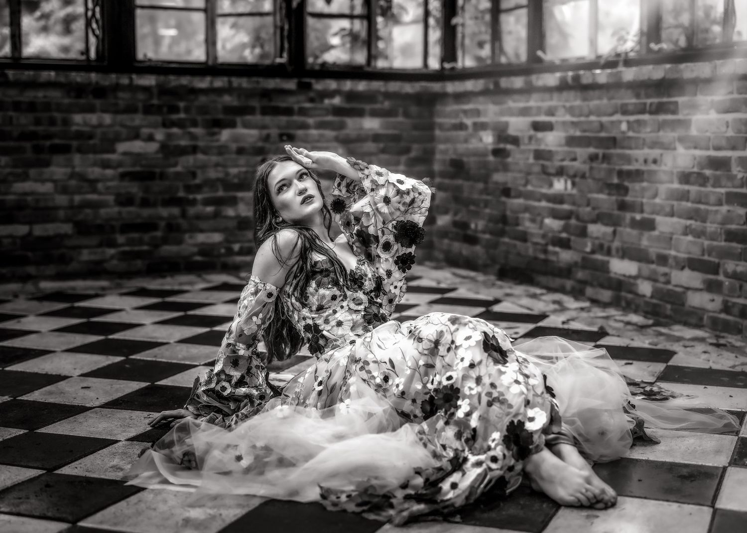 Black and white photo of a woman in a floral dress sitting gracefully on a checkered floor near brick walls.