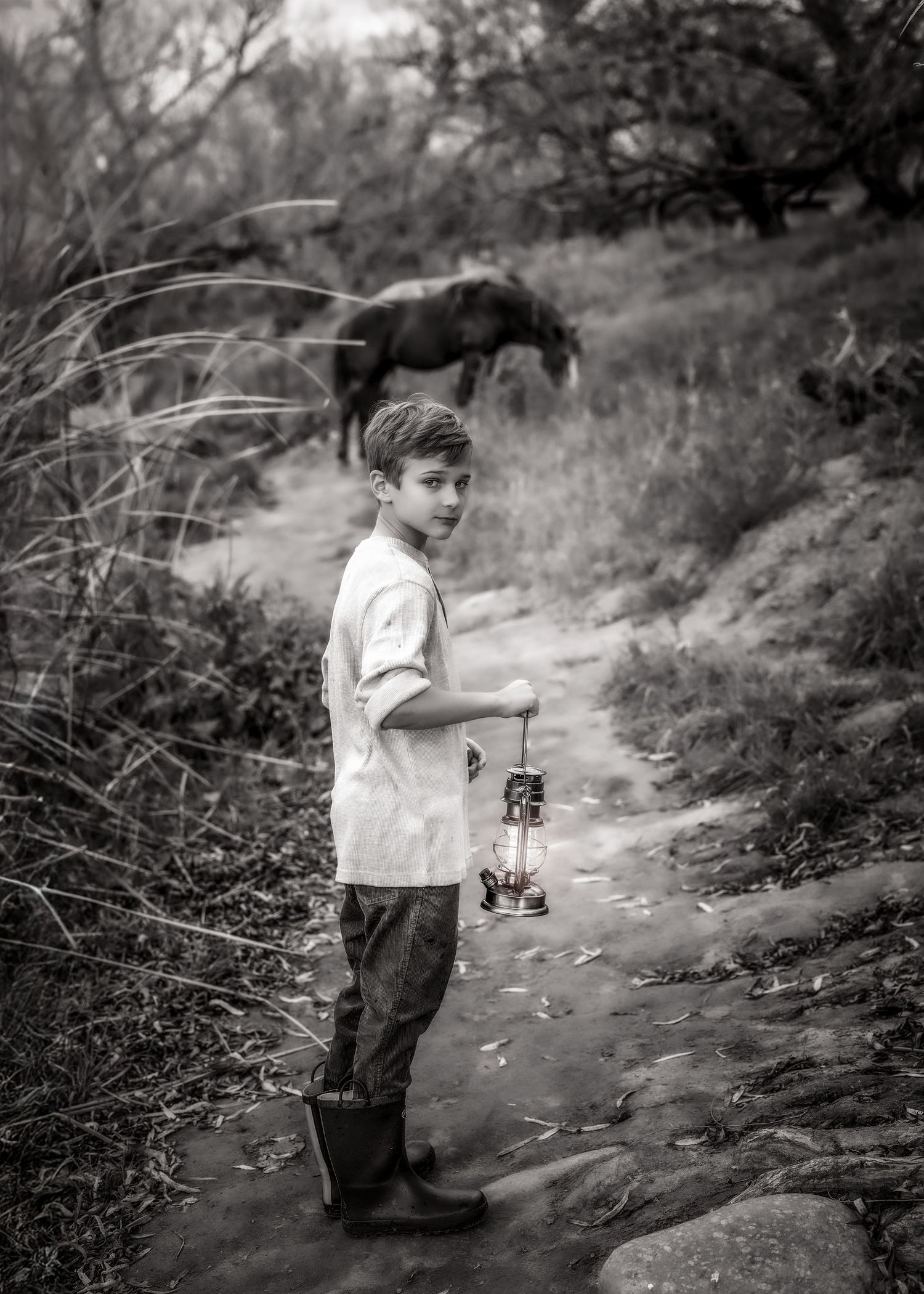Black and white photo of a boy holding a lantern on a forest path with a horse grazing behind.