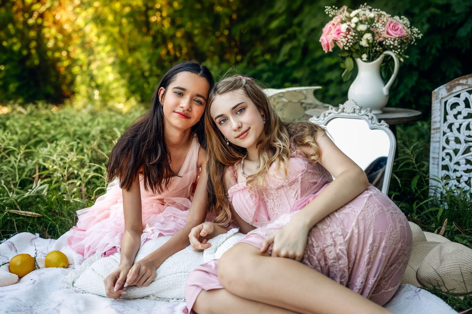 Two young women in pink dresses enjoy a picnic on a white blanket in a lush green garden with flowers.