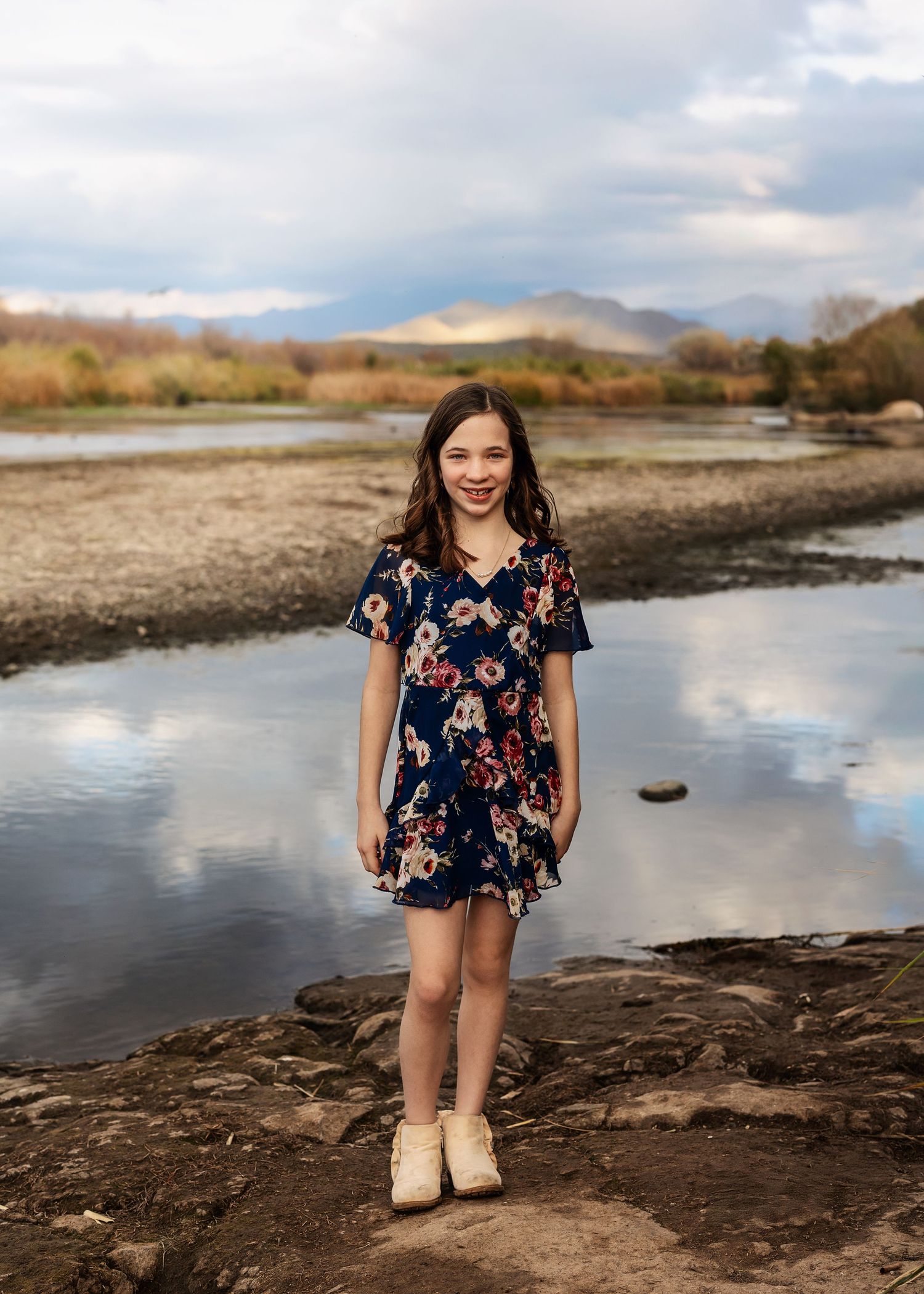 Smiling girl in floral navy dress stands on rocks beside a reflective river with mountain backdrop.