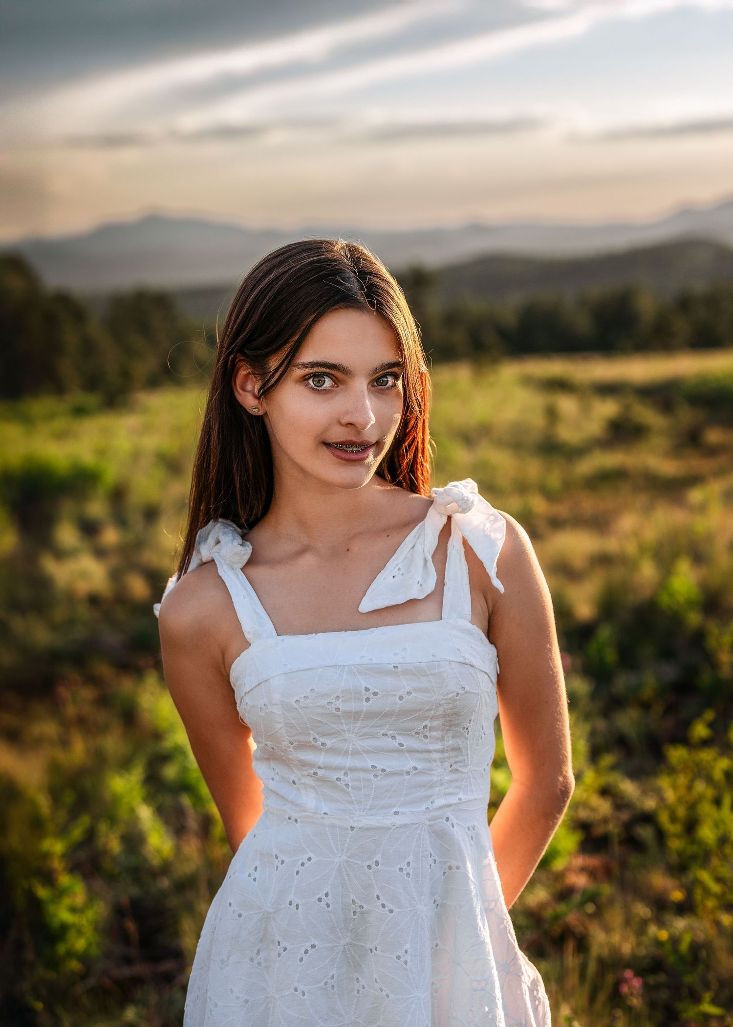 Young woman in white eyelet dress poses in a sunlit mountain meadow at golden hour.