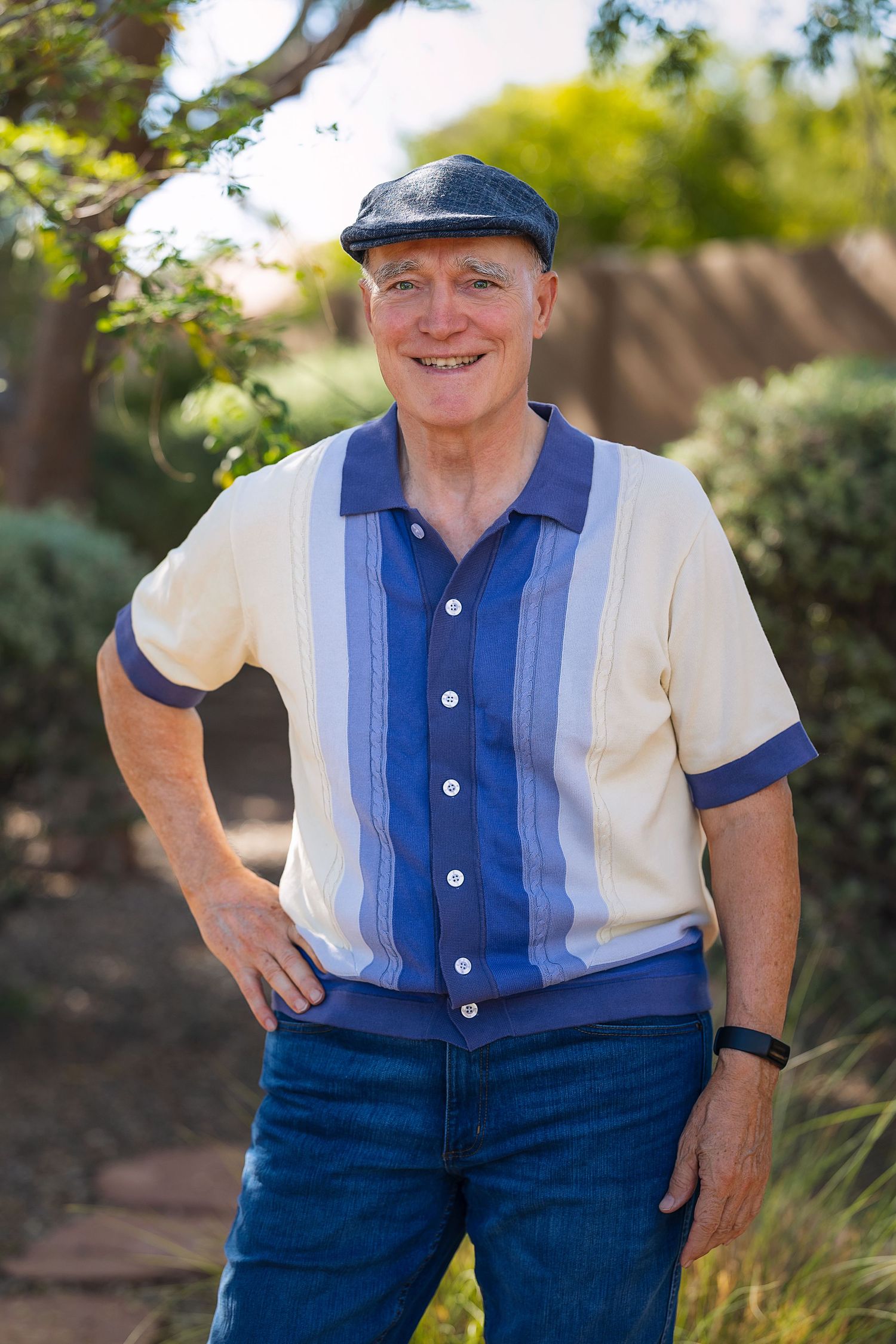 Smiling senior man in a blue striped polo shirt and flat cap posing with hands on hips outdoors.