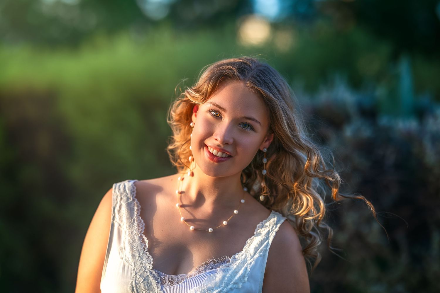 Smiling bride in white dress with pearl necklace, golden hour sunlight, lush green garden background.