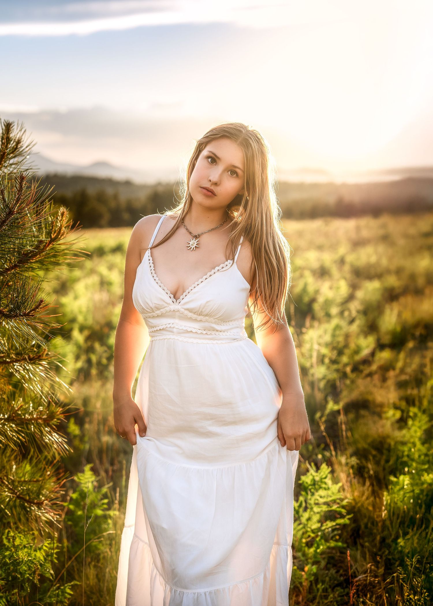 Blonde woman in white sundress standing in golden field at sunset with mountain backdrop.