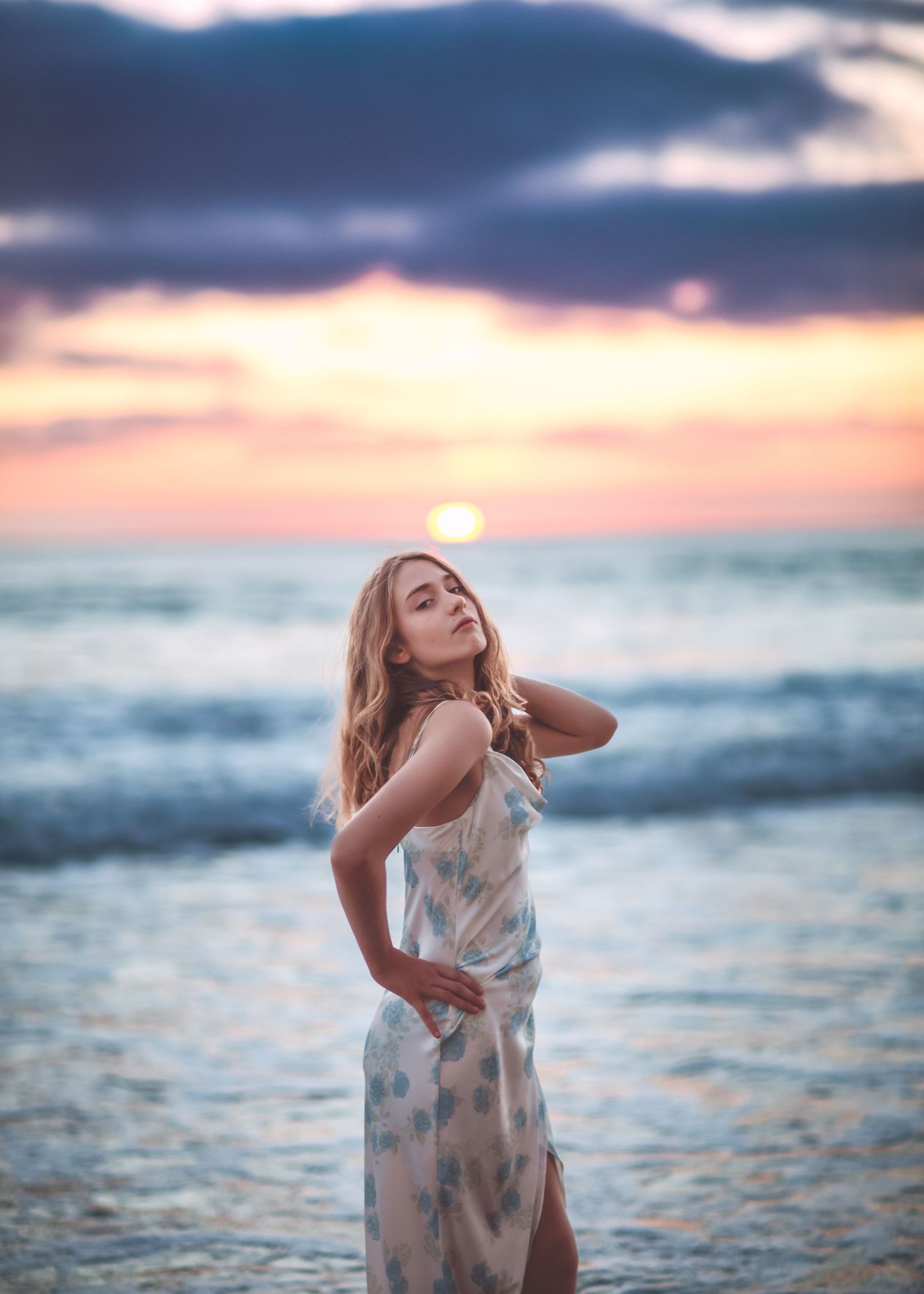 Blonde woman in floral dress posing on beach at dramatic sunset.