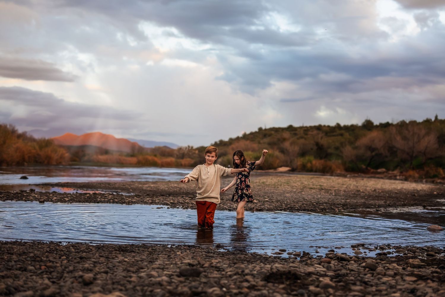 Two children laugh and play in a shallow river with colorful autumn landscape and dramatic clouds.