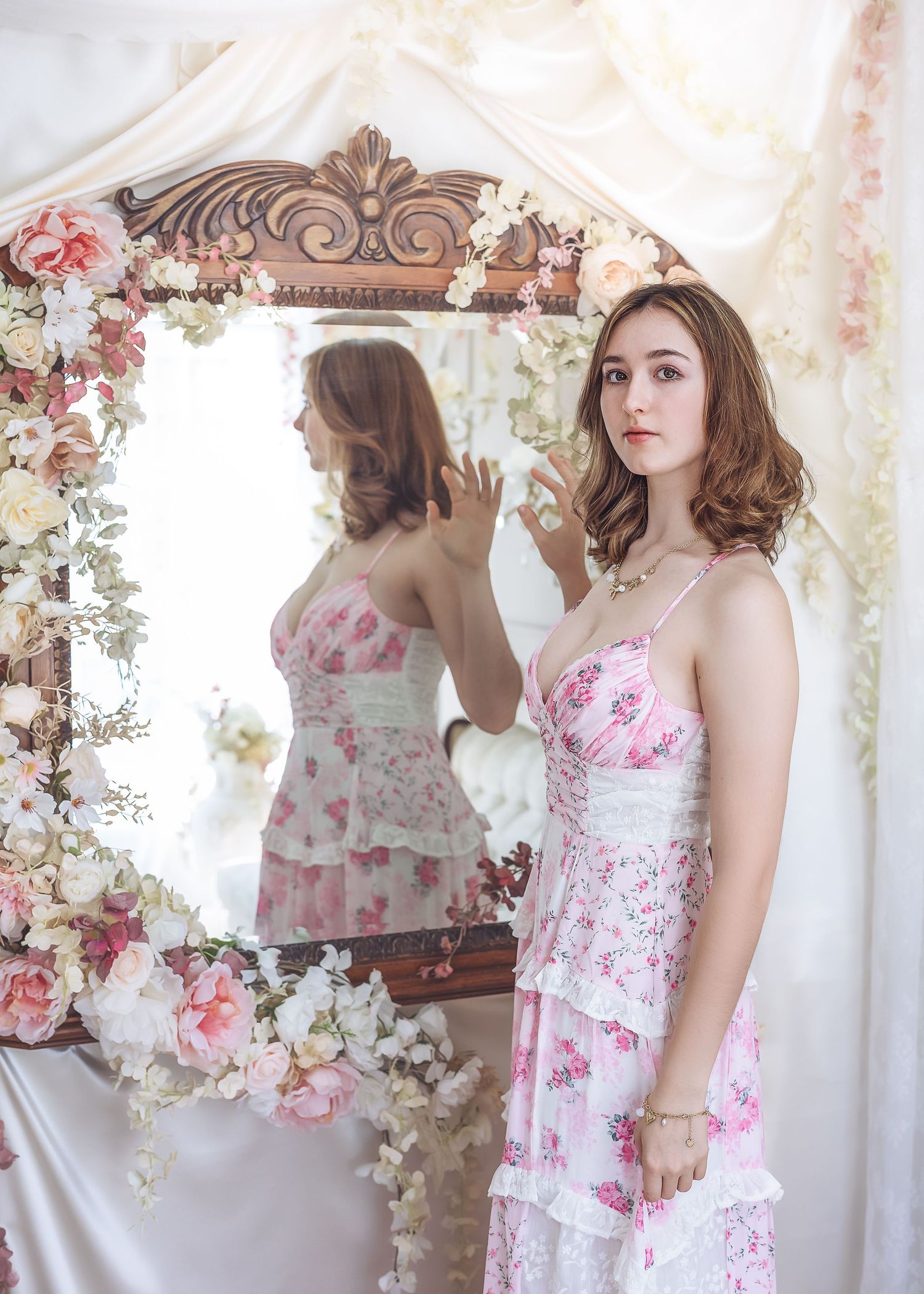 A young woman in a pink floral dress stands before a flower-adorned ornate mirror in soft lighting.