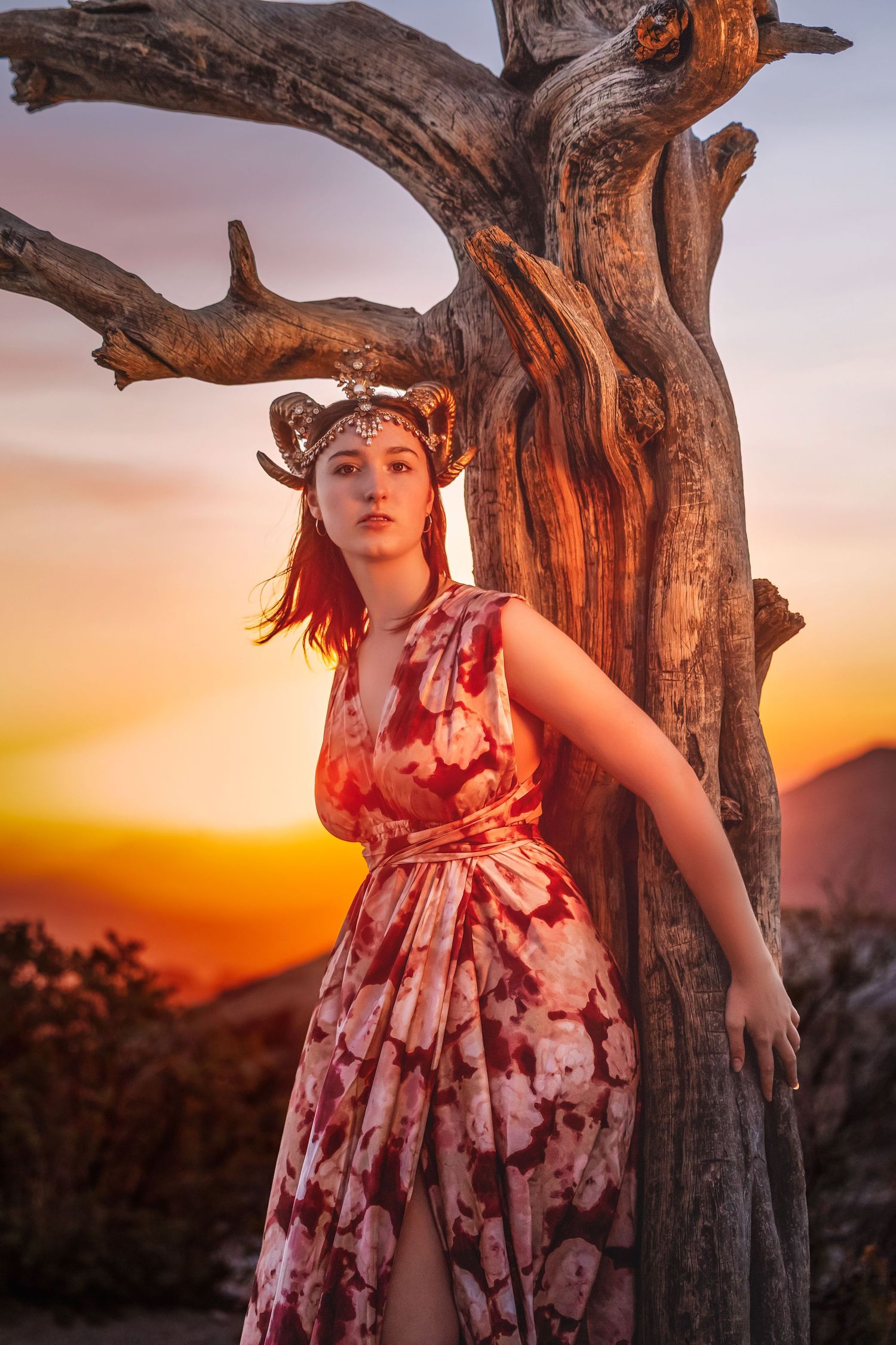 Woman in red floral dress with antler crown leans against dead tree at golden sunset.