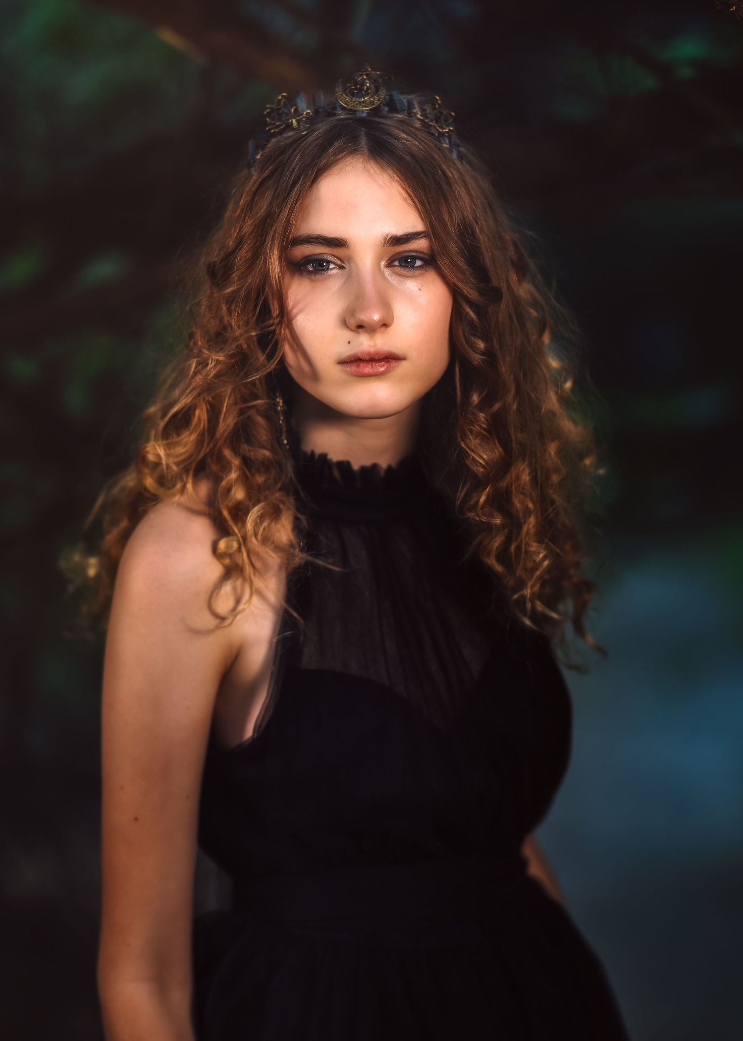Young woman with curly brown hair wearing a black dress and dark crown poses outdoors in soft forest light.