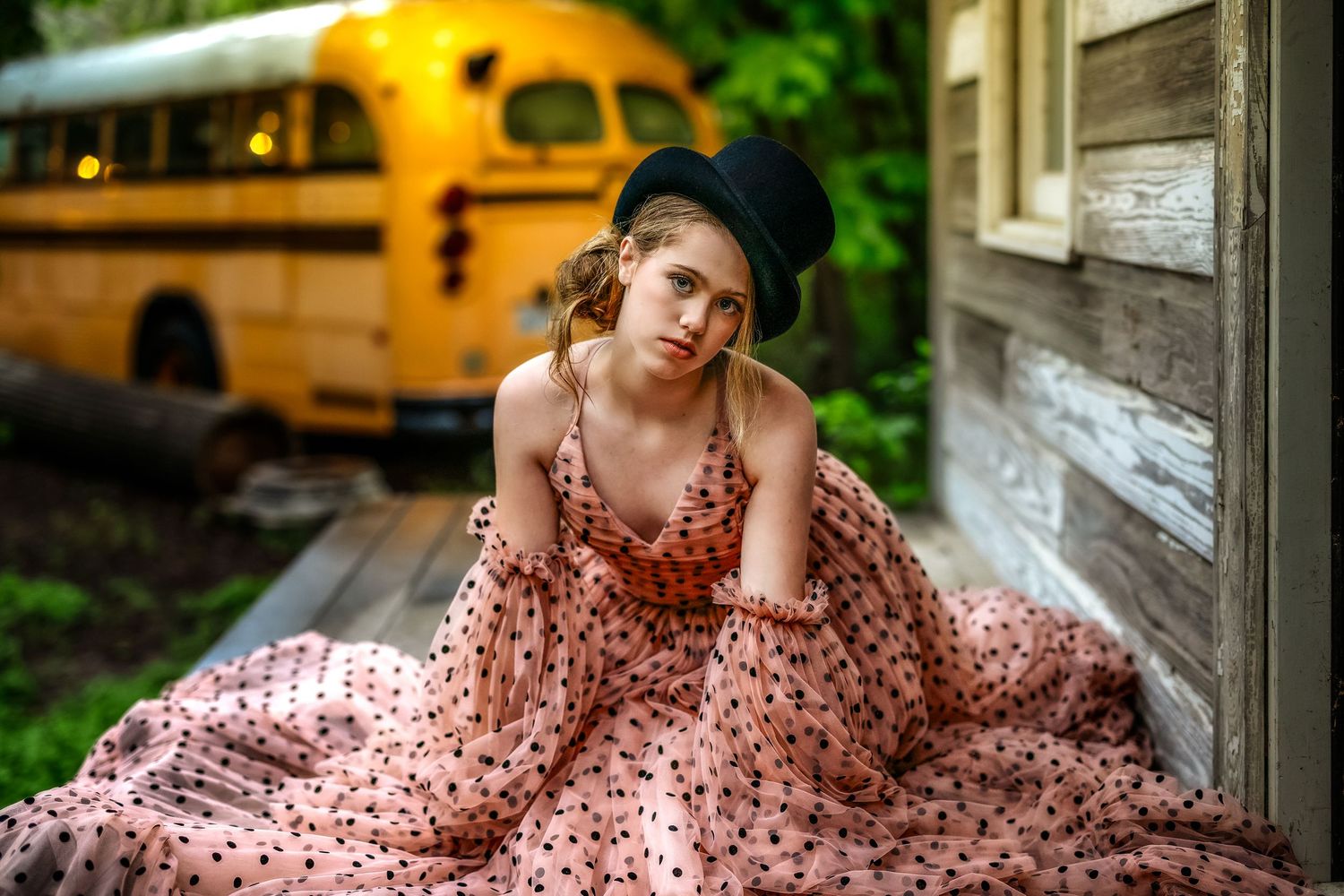 Young woman in pink polka dot dress and black top hat sits on porch steps with yellow school bus in background.