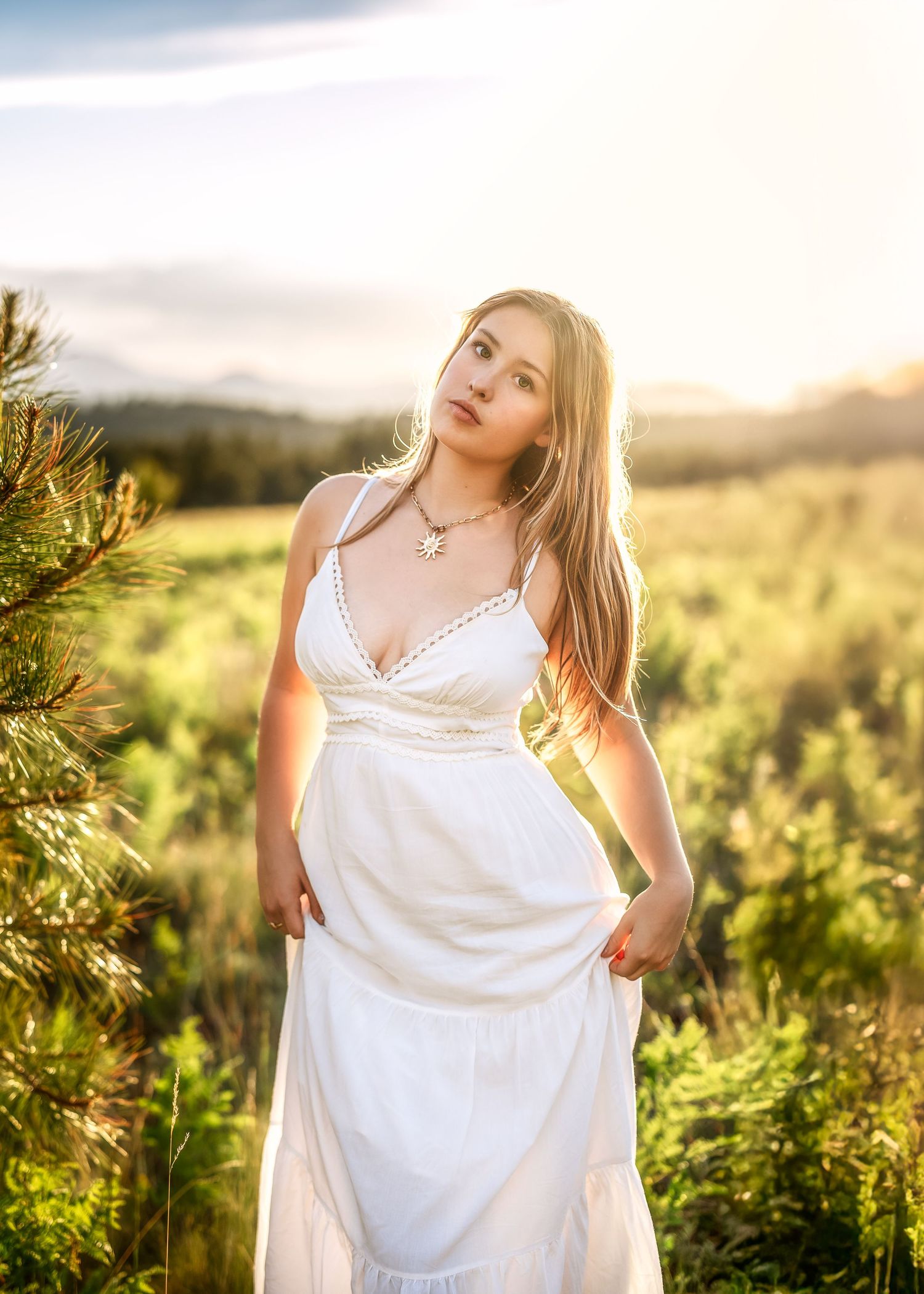 Young woman in white sundress standing in golden meadow with mountain sunset backdrop.