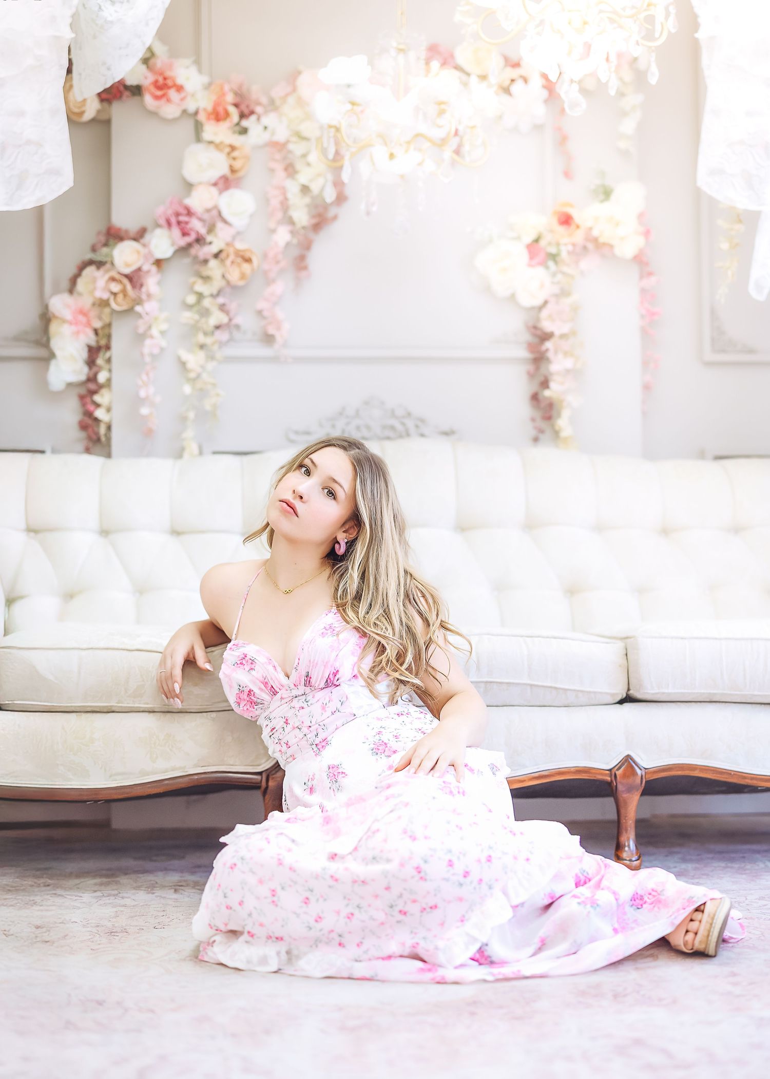 Young woman in white pink floral gown sitting beside tufted sofa gazing upward in floral studio.