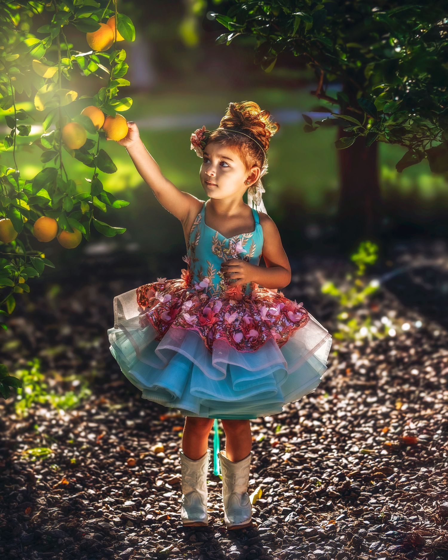 Little girl in colorful tutu dress reaching for oranges on a tree in a sunlit garden.