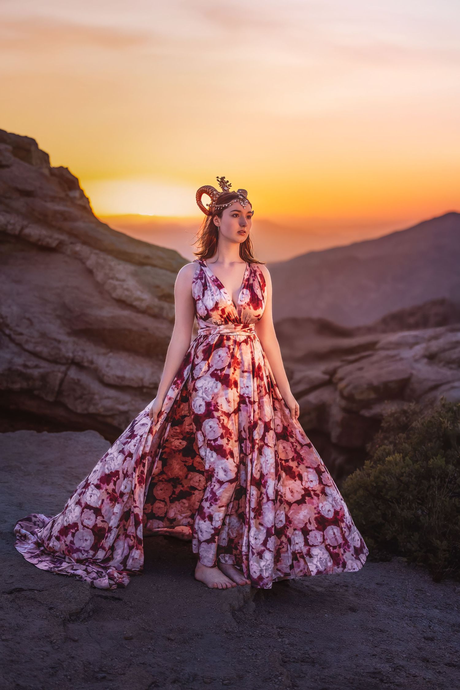 Woman in full floral gown with antler crown stands on rocks at glowing orange sunset.
