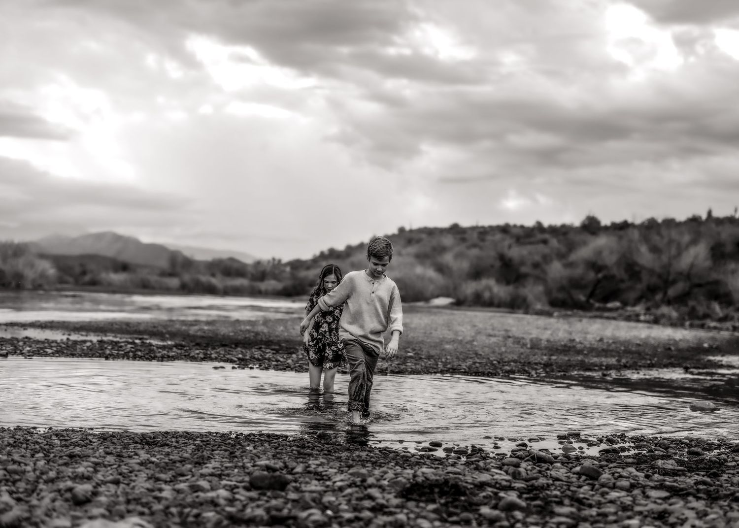 Black and white photo of two children wading together in a shallow rocky river under cloudy skies.