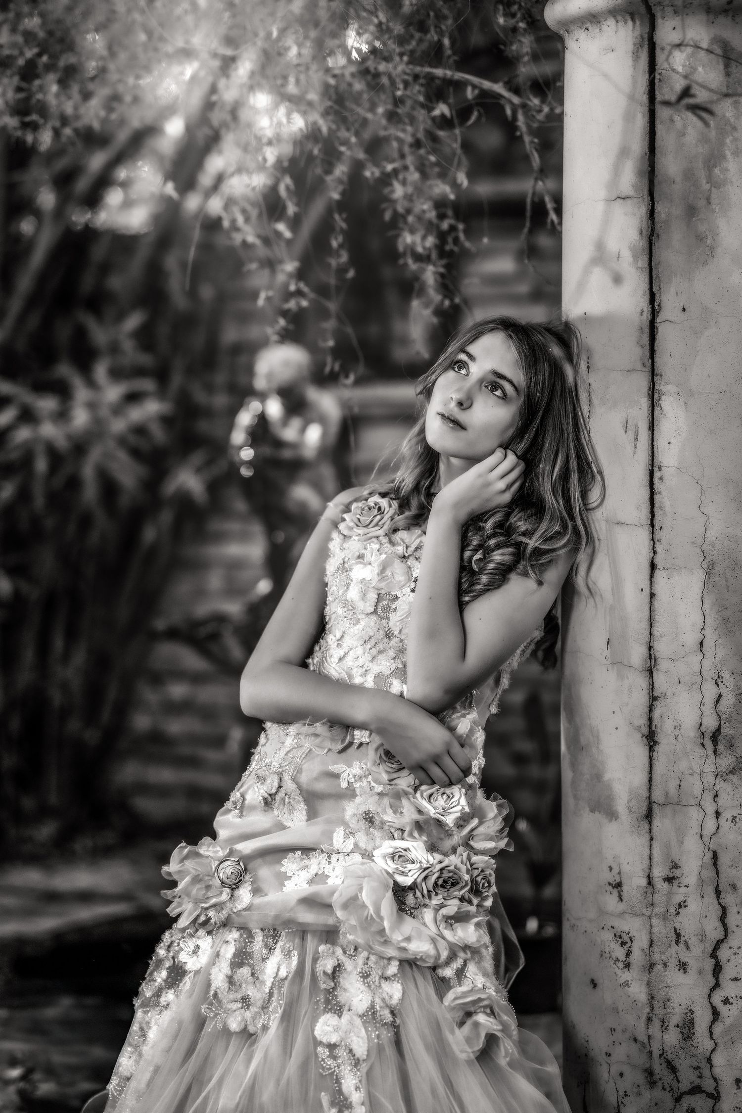 Black and white photo of young girl in floral dress leaning dreamily against rustic pillar.