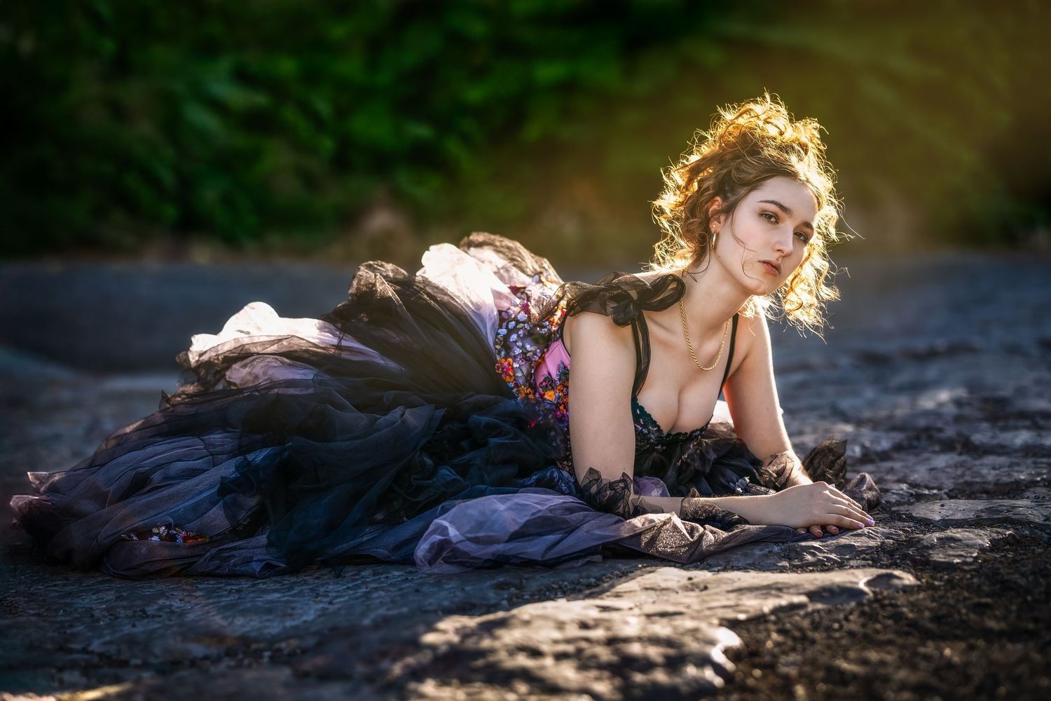 A woman in a dark flowing gown lies gracefully on rocky ground in soft golden sunlight.