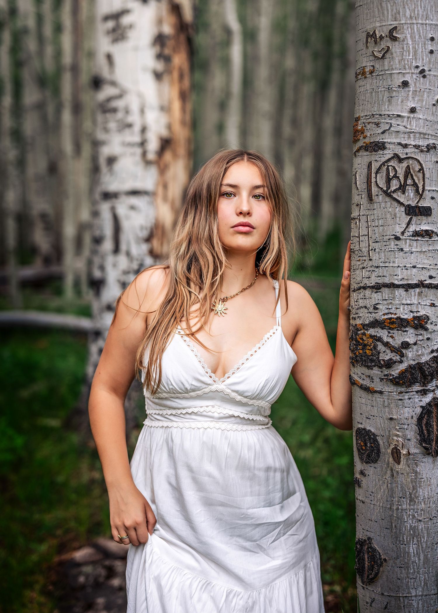 Young woman in white sundress leaning against birch tree in forest.