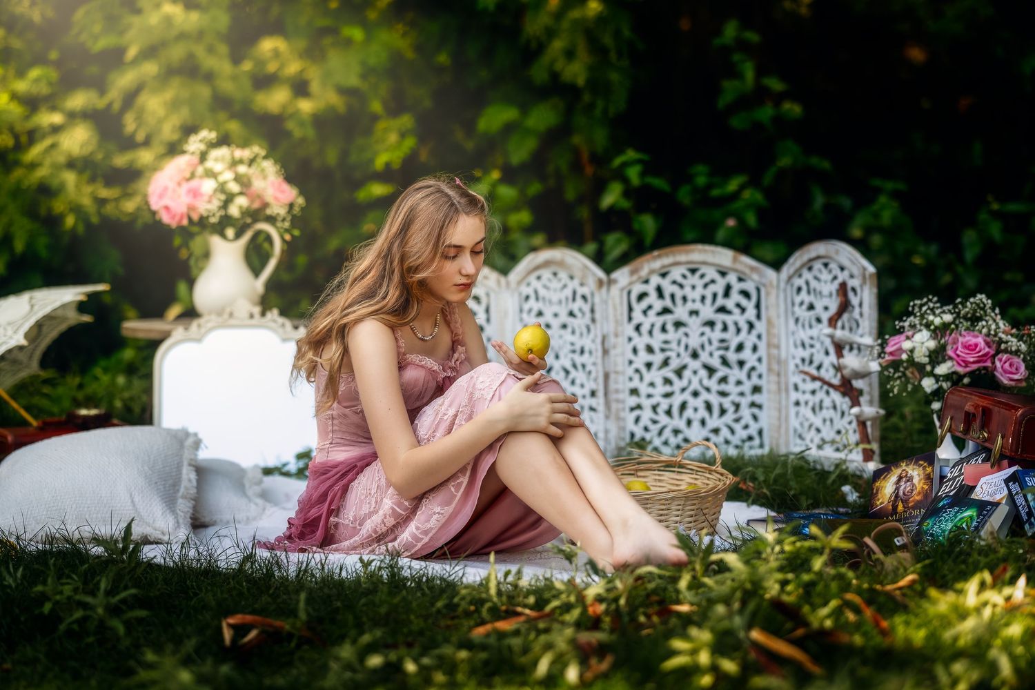 Young woman in pink dress sitting outdoors in garden, holding an apple near decorative white screen.