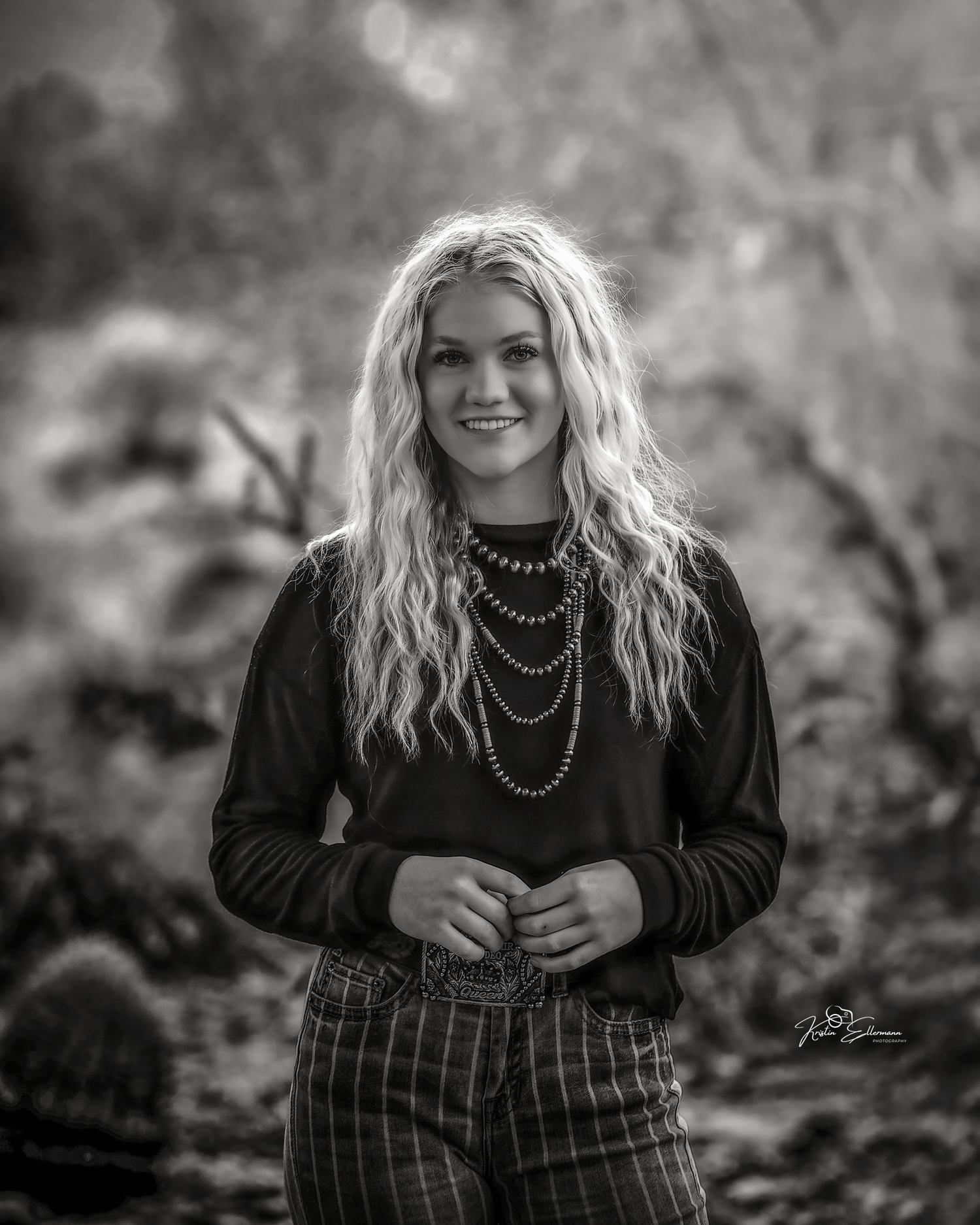 Black and white portrait of smiling blonde teen girl in black sweater and layered necklaces in desert setting.