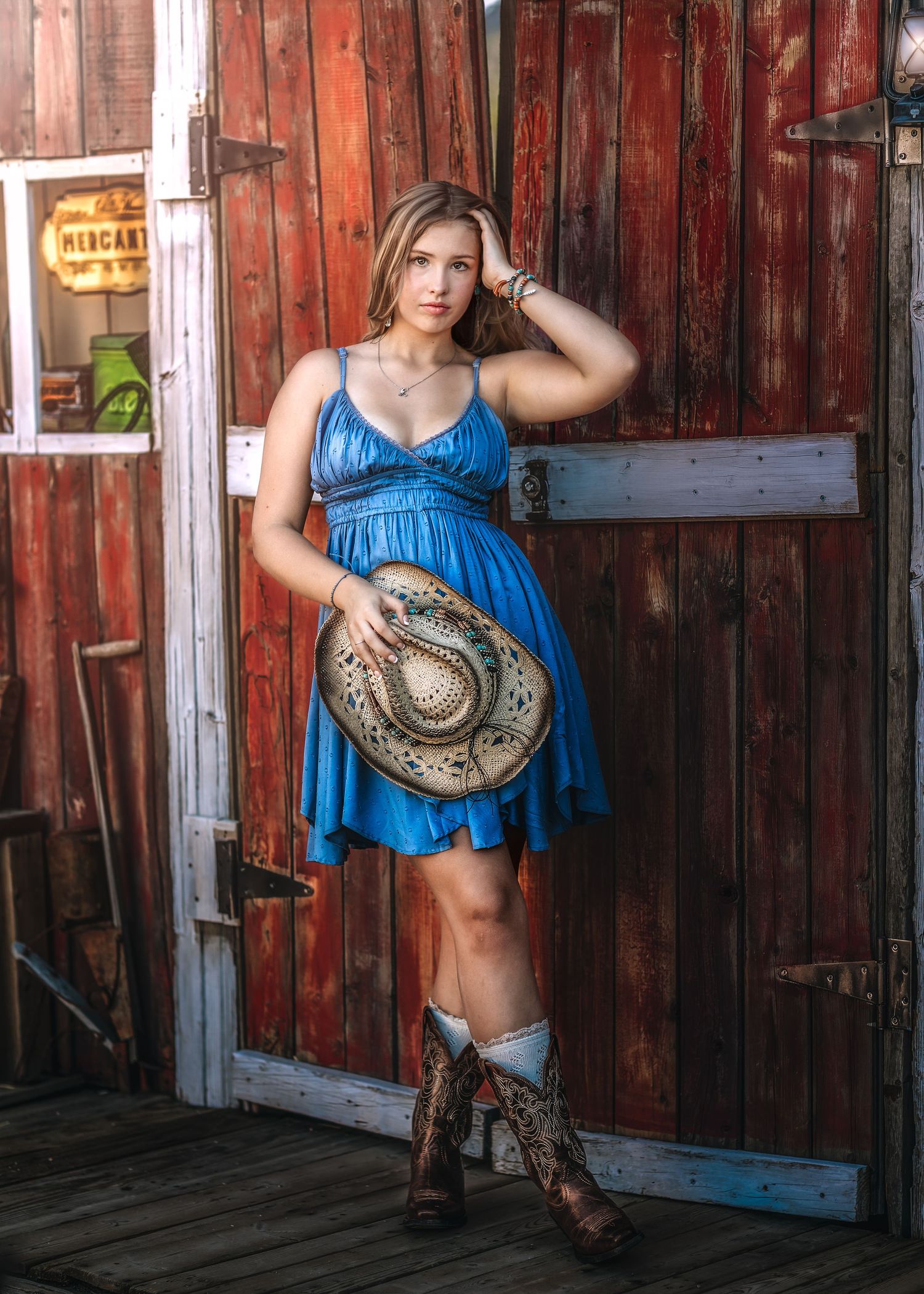 Young blonde woman in blue dress holding cowboy hat leaning against red barn door in boots.