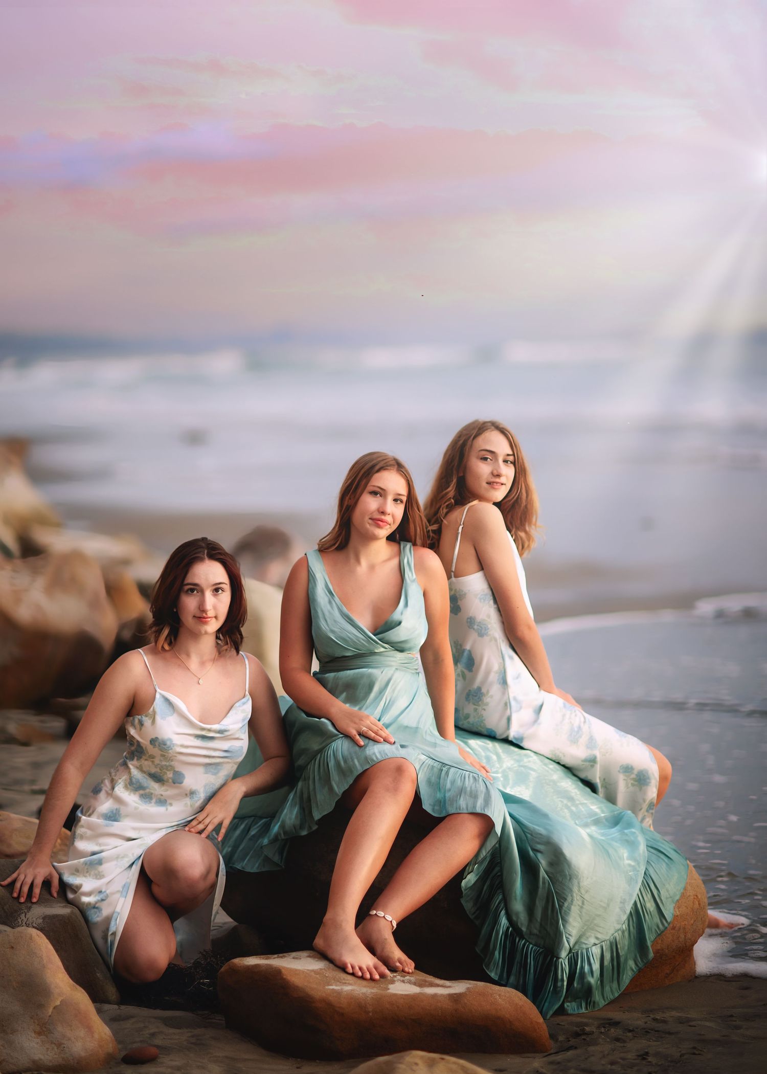 Three young women in elegant dresses pose on beach rocks under vibrant pink and purple sunset.