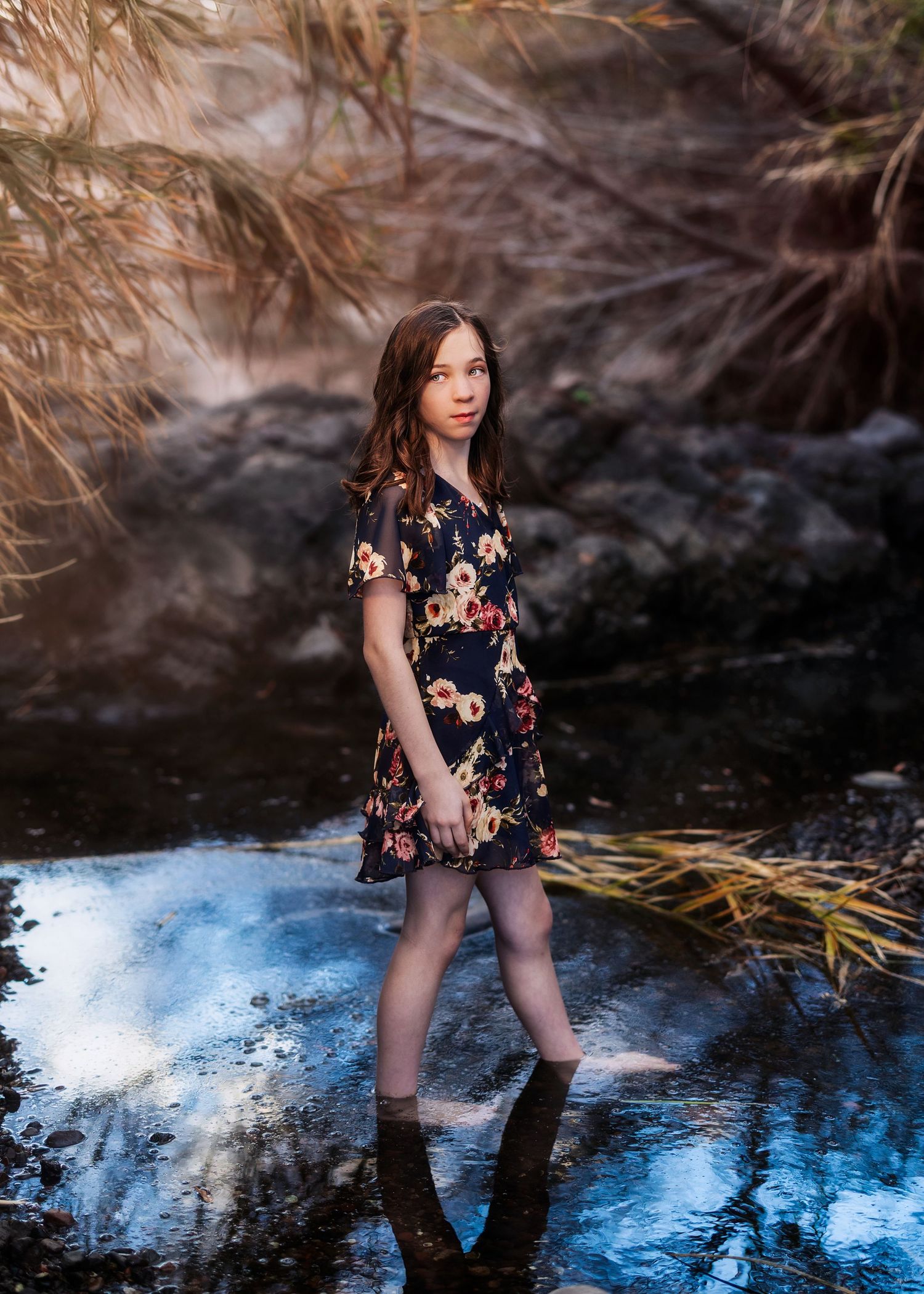 Girl in dark floral dress standing in shallow water surrounded by reeds and rocks.