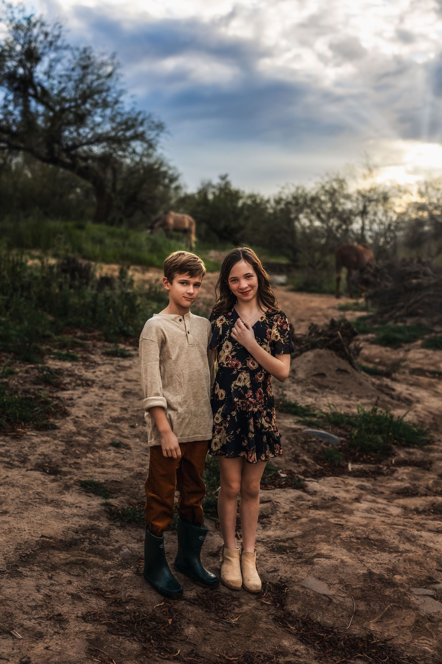 A boy and girl stand together on a dirt path at sunset with horses grazing in the background.