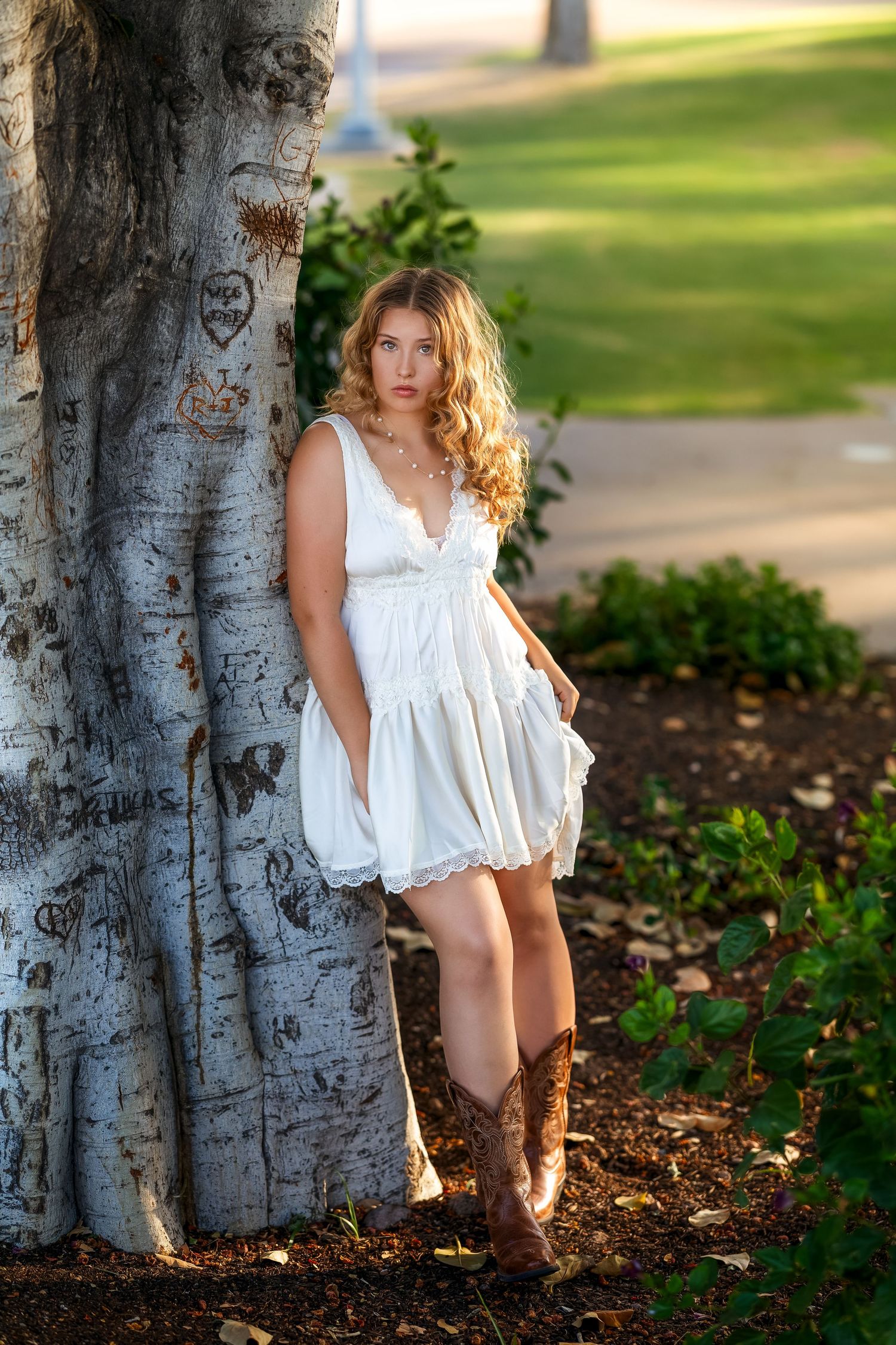 Young woman in white dress and cowboy boots leaning against a carved tree in a lush green park setting.
