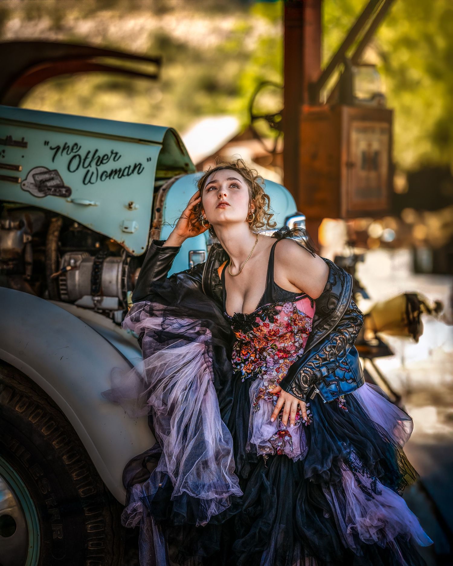 Woman in ornate gown leans against vintage truck labeled The Other Woman in rustic outdoor setting.