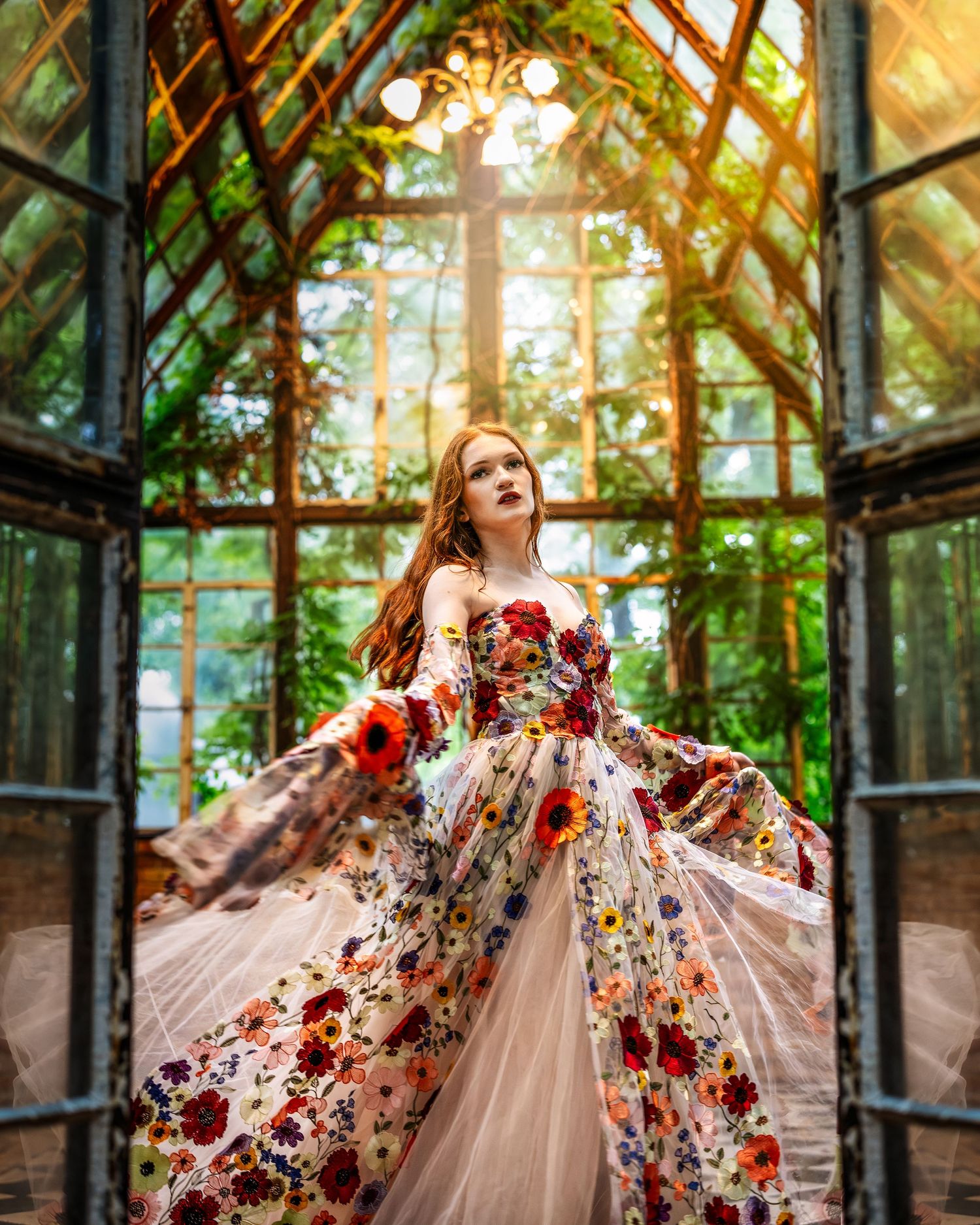 Woman in floral gown twirling in sunlit greenhouse with lush greenery and vintage iron frame windows.