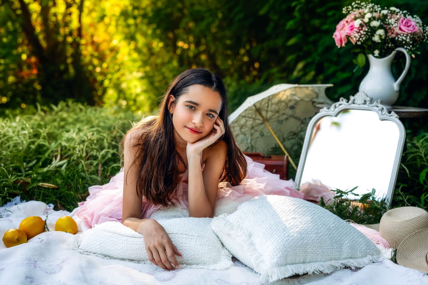 A young woman in a pink dress relaxes on white pillows outdoors near a mirror and floral arrangement.