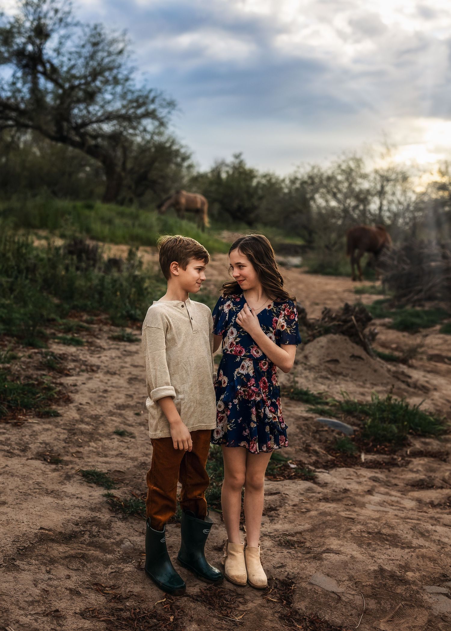 A boy and girl smile at each other on a dirt path with horses grazing behind them.