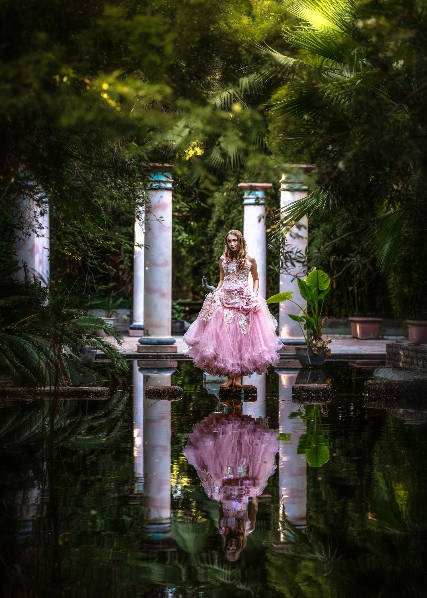 Woman in pink ball gown reflected in still water surrounded by lush greenery and white columns.