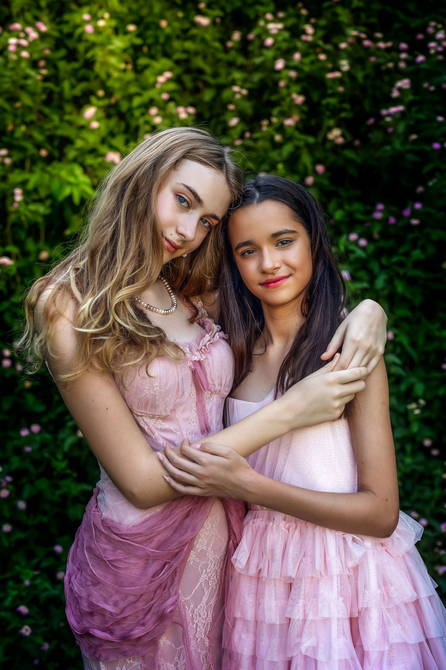Two girls in pink tulle dresses embrace in a garden with green foliage and pink flowers blooming behind them.