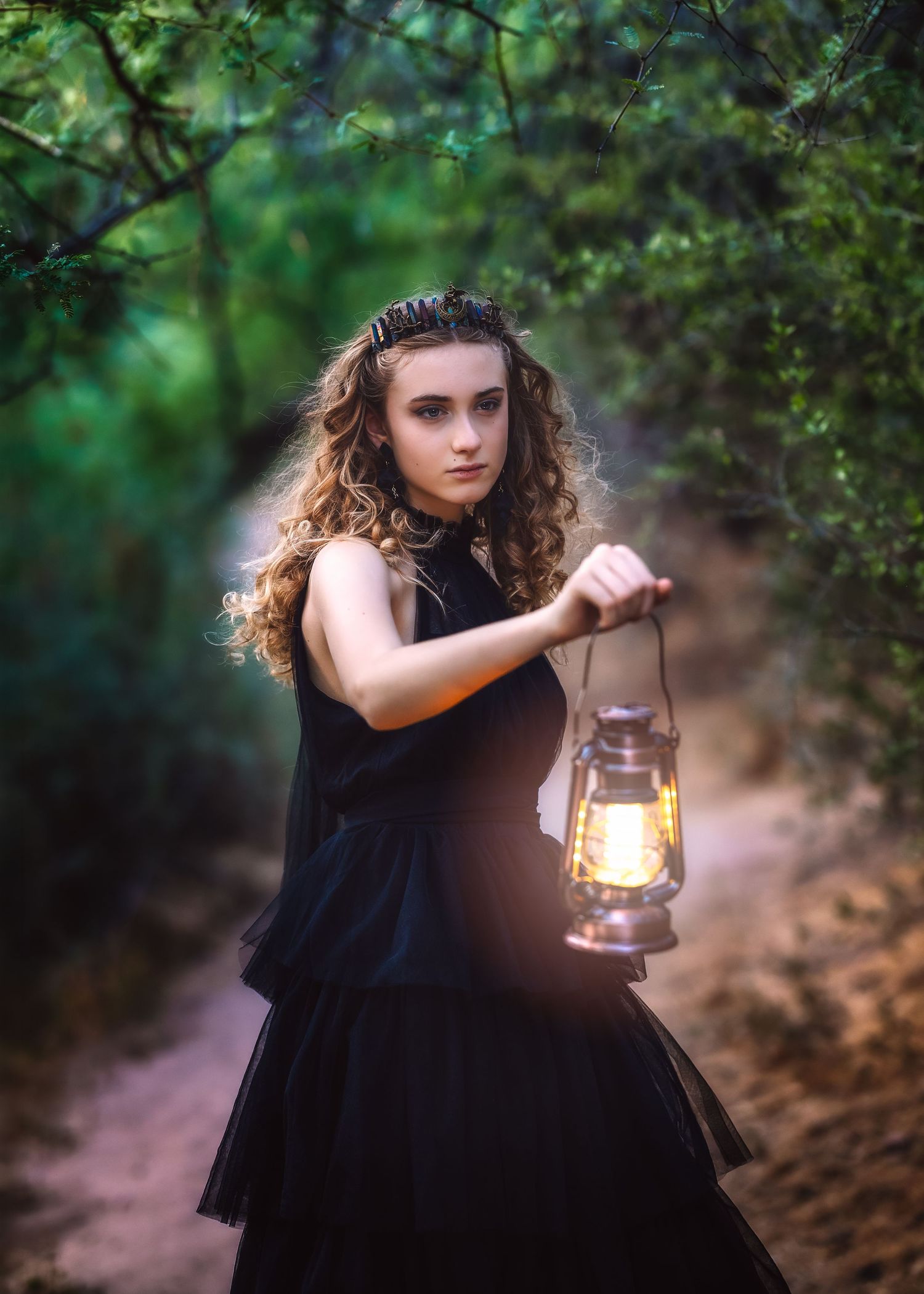 Young woman in black dress holding a glowing lantern on a forest path at dusk.
