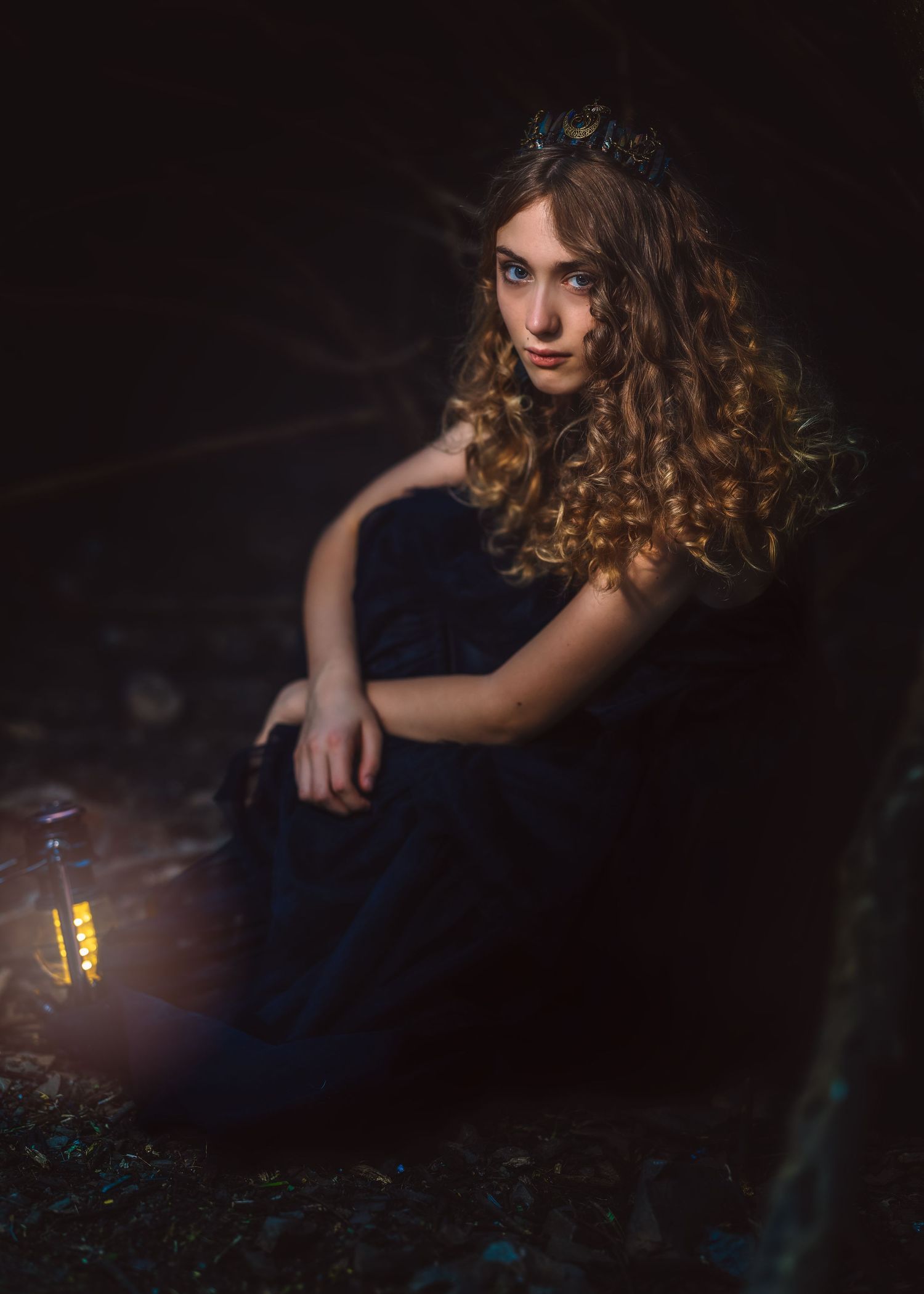 Young woman in dark blue gown with curly hair and floral crown sits beside a glowing lantern in a dark forest.