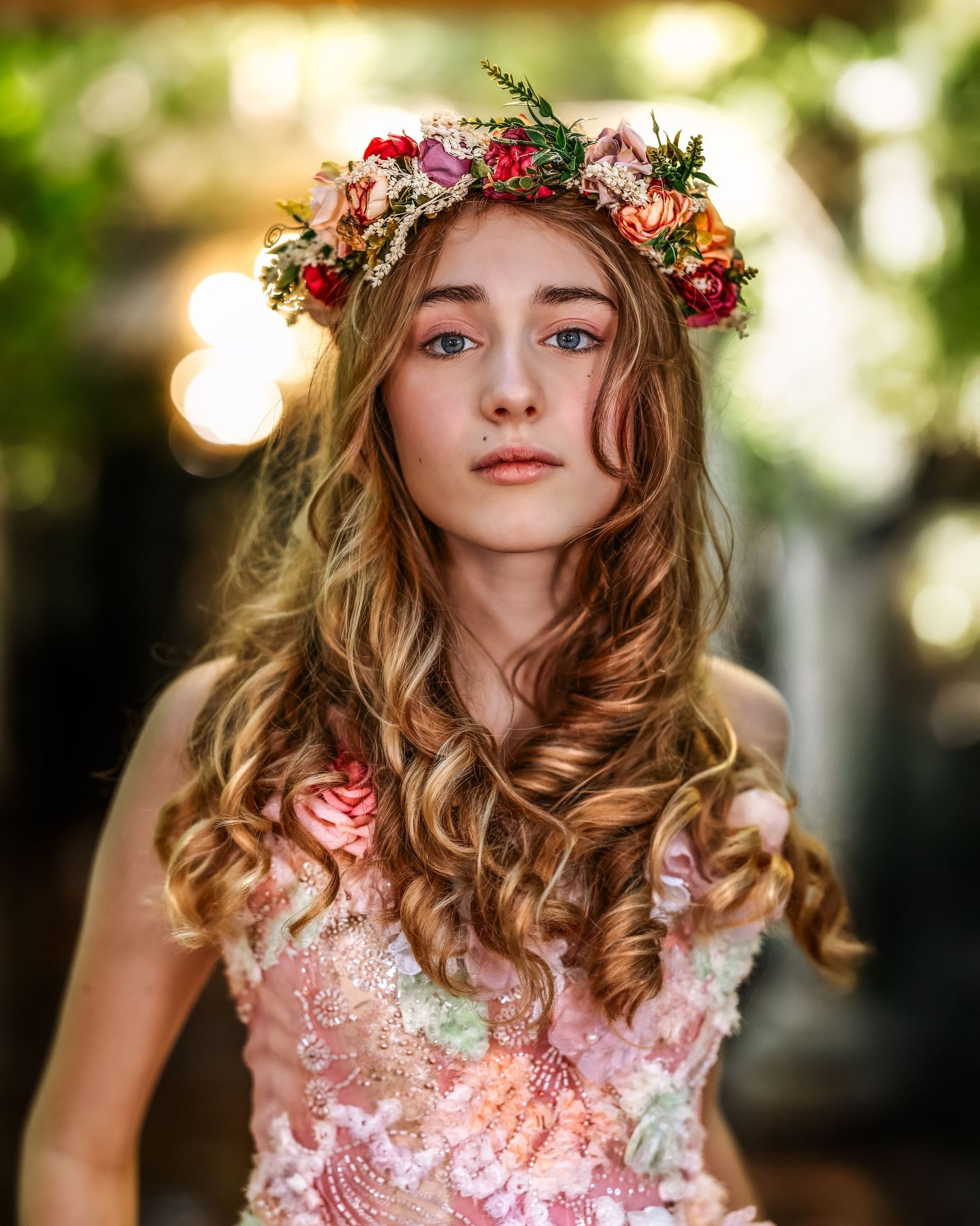 Young woman with long wavy hair wearing a colorful floral crown and pink embroidered dress, posed outdoors.