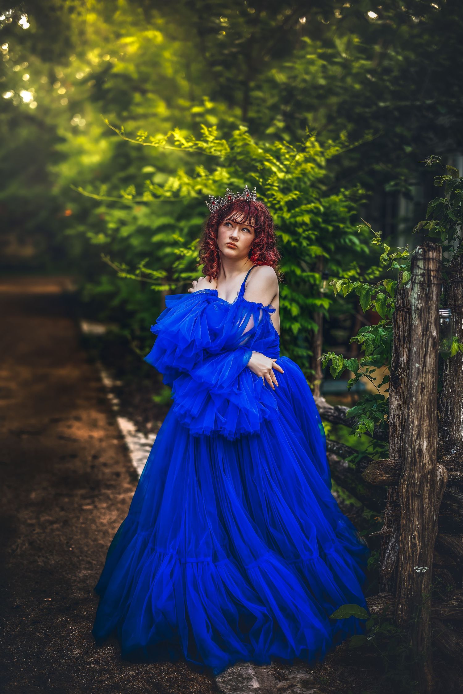 Woman in royal blue ruffled ball gown with crown poses in lush green garden pathway.
