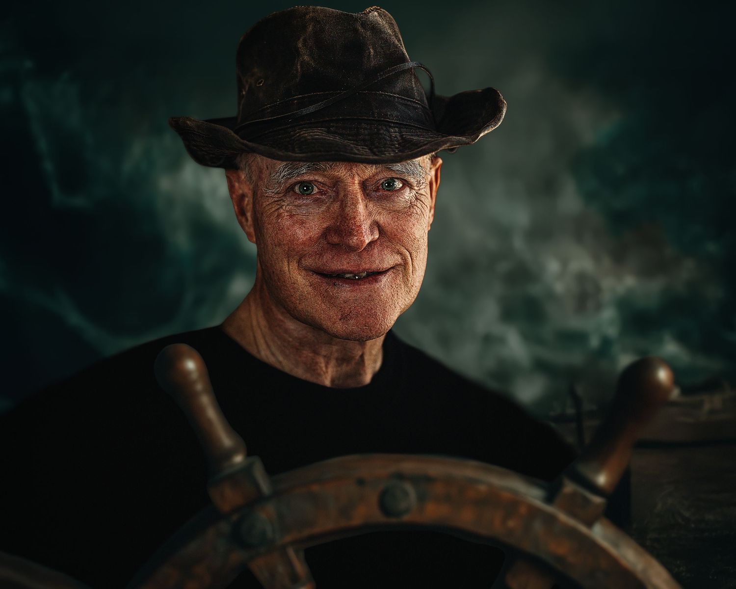Elderly man in a weathered hat smiling at the helm of a ship's wheel against a dramatic stormy background.
