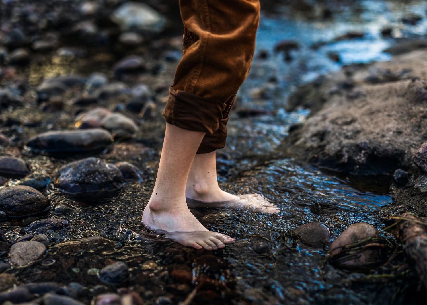 Barefoot child standing in shallow rocky stream with brown pants rolled up.