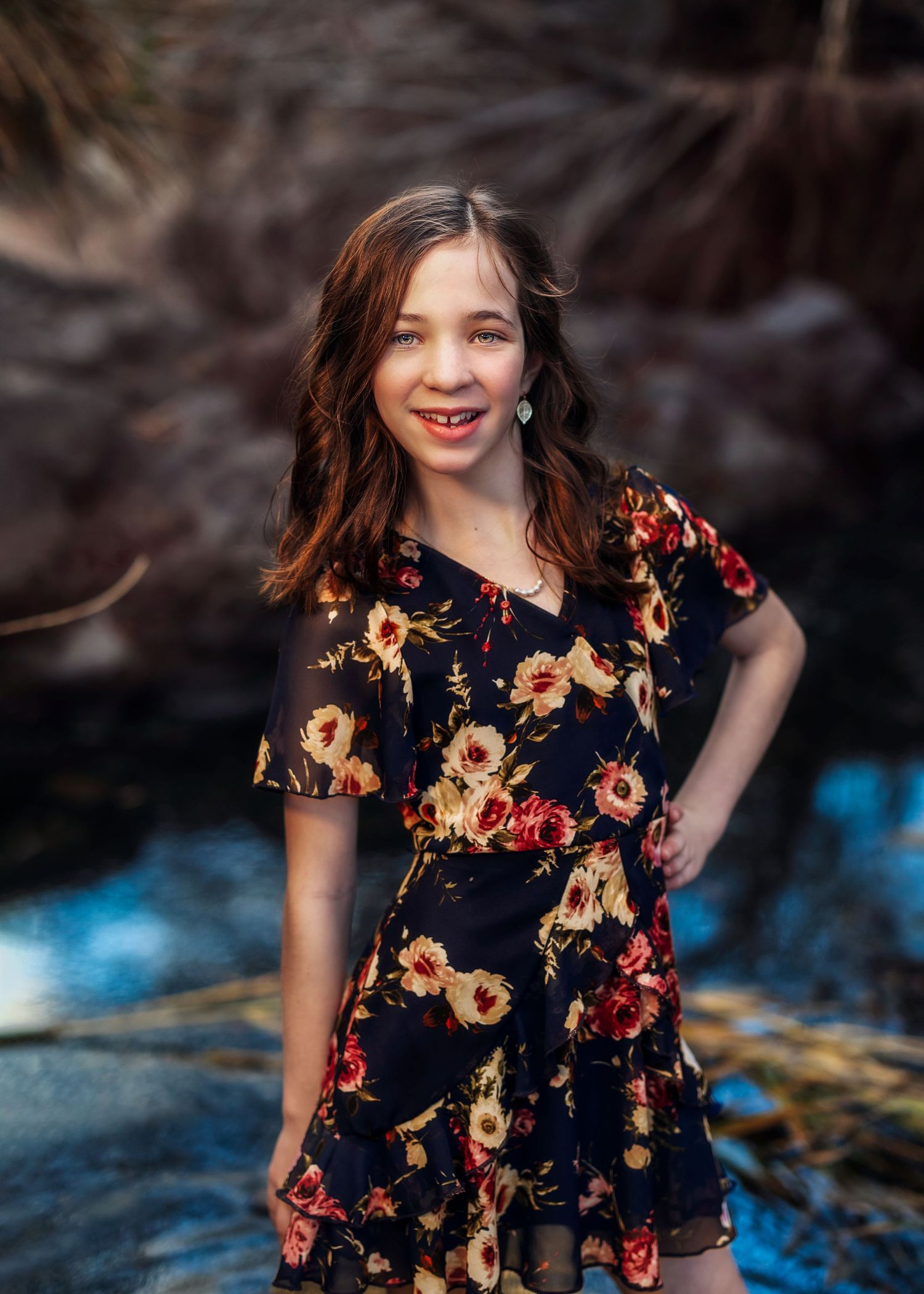 Smiling girl in dark floral dress posing outdoors near rocky background in natural light.