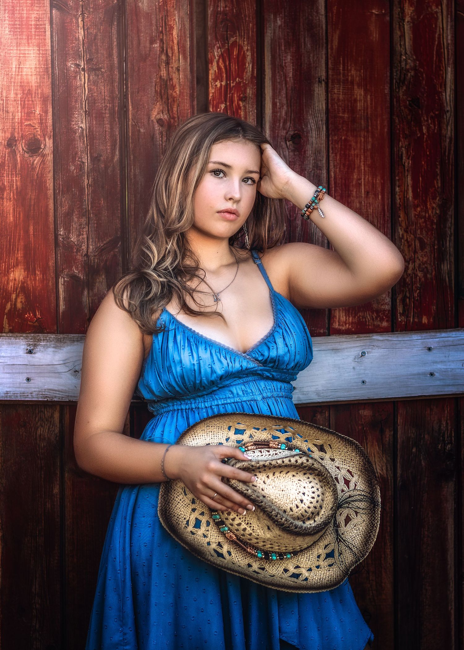 Young woman in blue dress holding decorated cowboy hat against rustic red wooden barn wall.