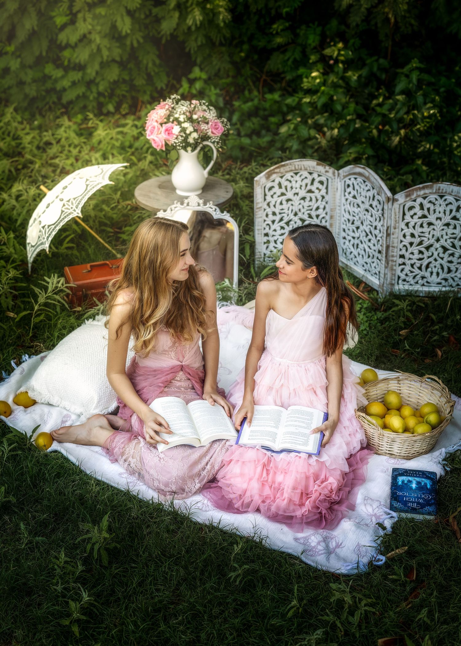 Two women in pink dresses sit on a blanket outdoors, reading books near lemons, flowers, and a decorative screen.