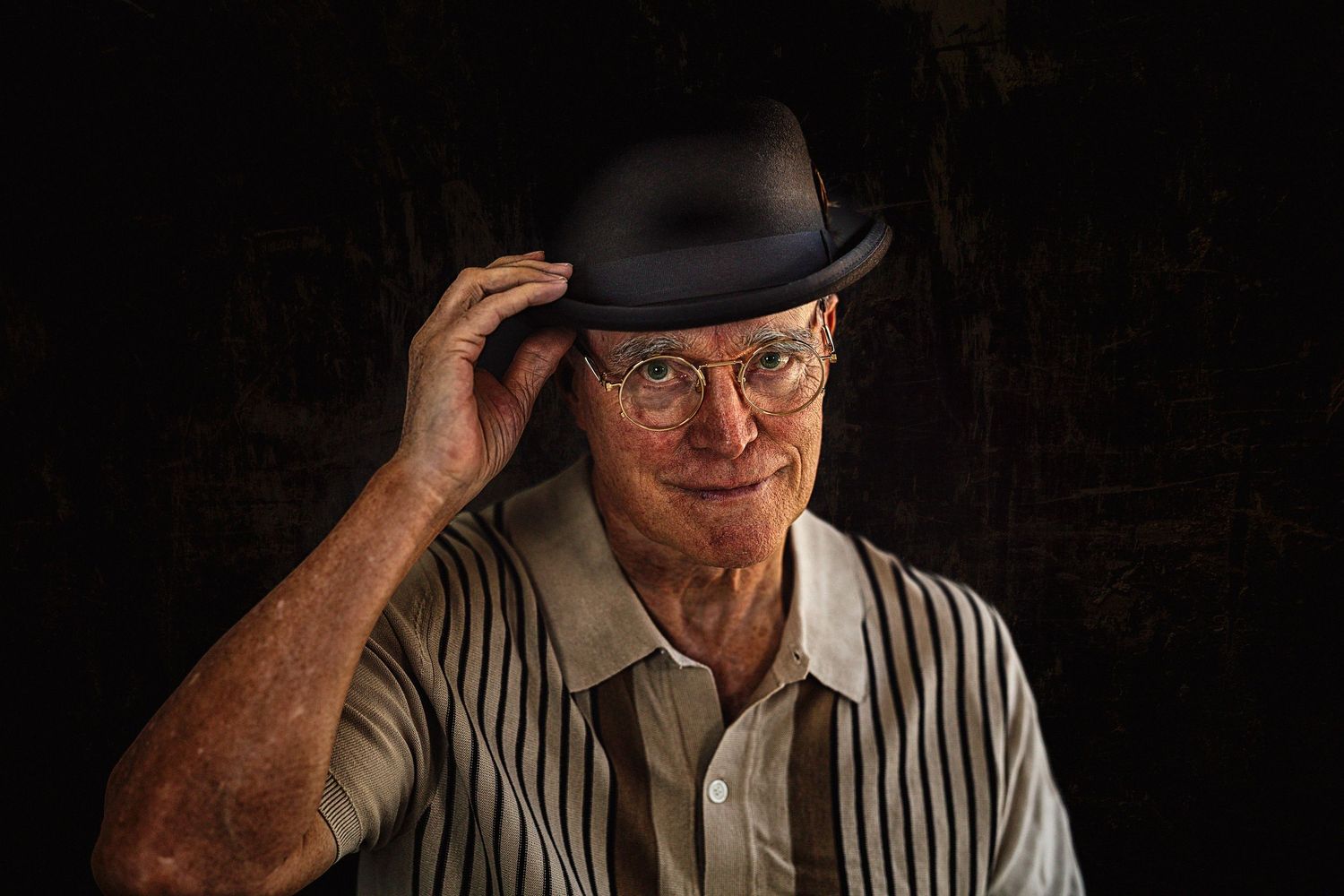 Older man tipping his black bowler hat while smiling, wearing striped shirt and glasses.